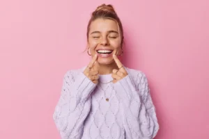Person with closed eyes pointing to a broad smile while standing against a pink background.